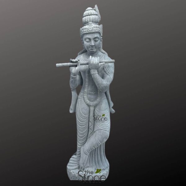 tall, slender, hand-carved grey natural stone statue of Lord Krishna is depicted in his iconic Tribhanga pose, gracefully playing the bansuri flute. The deity stands with one leg bent at the knee and crossed over the other, creating a dynamic yet serene posture. His hands are delicately positioned to hold the horizontal flute (bansuri) to his lips, with fingers poised as if producing a melodic tune. Krishna's face exudes a peaceful, meditative expression, with eyes gently downcast, full lips, and a subtle, soft smile. He wears an ornate, multi-tiered crown adorned with intricate carvings and a distinctive plume-like element at the top, subtly suggesting a stylized peacock feather. Elaborate, traditional Hindu earrings grace his elongated earlobes, and finely sculpted strands of hair flow down his shoulders. His slender, youthful body is richly adorned with finely chiseled jewelry, including multiple necklaces (malas) of varying lengths, intricately detailed armlets on his upper arms, and bracelets on his wrists. A sacred thread (yajnopavita) is subtly visible across his torso. His lower body is draped in a flowing dhoti-like garment, characterized by deep, realistic pleats and folds that showcase the artisan's skill in stone carving, and secured by an ornate waist belt. Anklets are visible around his bare feet. The dark grey natural stone has a slightly textured, unpolished finish, highlighting the raw beauty of the material and giving the sculpture an ancient, timeless quality. The entire statue stands on a simple, round base. The background is a solid, dark grey, ensuring the exquisite details and masterful craftsmanship of this Hindu deity statue stand out prominently. This exquisitely crafted murti serves as a powerful piece of traditional Indian spiritual art, suitable for home altars, meditation spaces, temples, or as an elegant example of cultural decor and religious sculpture.