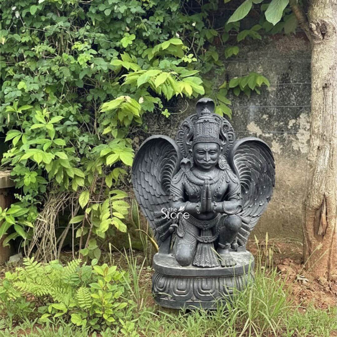 3 feet Garuda Statue 3 A powerful 3-foot Blackstone Garuda statue serving as a striking Garuda Statue for Garden Decor is prominently situated in a verdant outdoor setting nestled against a lush green vine-covered wall and a tree trunk. The statue carved from deep lustrous black stone depicts Garuda the mythical eagle-like deity and vehicle of Lord Vishnu in a reverent kneeling or crouching posture. His hands are pressed together in Anjali Mudra signifying devotion and respect. This posture combined with his formidable features and outstretched wings makes him a captivating presence. Garuda is depicted with a human-like torso and legs a majestic bird-like face with a beak-like nose and large intricately carved wings spread behind him ready for flight. He wears an elaborate crown adorned with a cobra traditional jewelry including necklaces and armlets and a sacred thread The intricate details of his feathers his powerful limbs and his facial expression which blends ferocity with devotion are all meticulously rendered in the black stone. The black stone's smooth finish highlights these fine details creating a striking contrast with the natural surroundings. The statue rests on a beautifully sculpted lotus-petal base also carved from black stone which provides a stable and dignified foundation. The surrounding environment is rich with various green plants including ferns and grasses creating a natural and harmonious backdrop. The textured tree trunk to the right and the dense green vines on the wall behind further integrate the statue into the garden landscape. This 3-foot Blackstone Garuda statue is an exquisite example of religious artistry designed to bring blessings protection and aesthetic grandeur to any garden or outdoor space perfectly aligning with an article focusing on such a revered and majestic Garuda statue for garden decor.