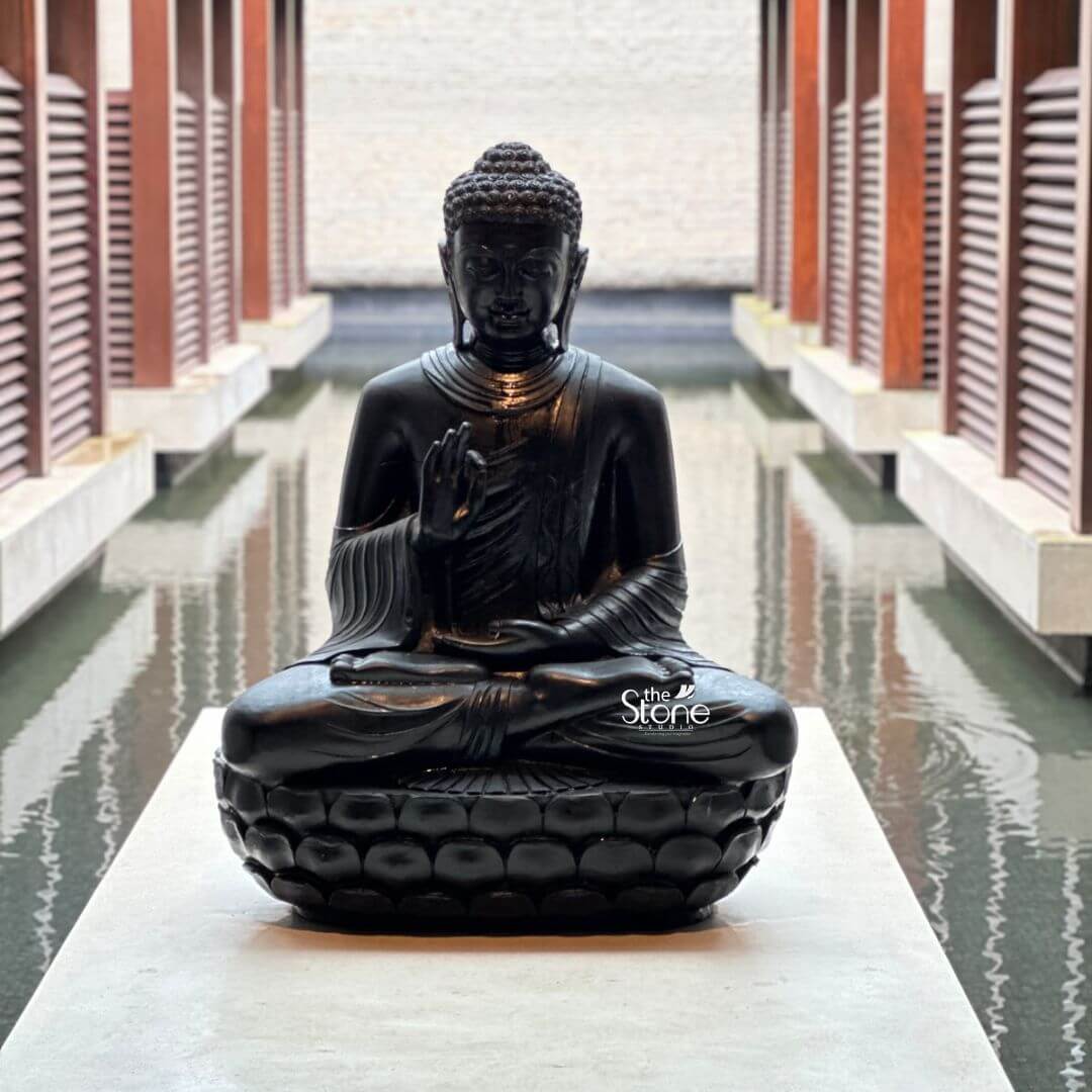 A centered eye-level photograph captures a serene four-foot-tall black stone Buddha statue seated in a meditative posture upon a raised white concrete plinth. The statue is positioned as the focal point within a tranquil open-air courtyard reflection pool at the Grand Hyatt Goa. The dark polished granite of the Buddha exhibits a smooth texture contrasting beautifully with the light stone of the plinth and the surrounding architecture. Lord Buddha is depicted with a calm compassionate expression gently closed eyes and tightly curled hair in the traditional ushnisha. His hands are held in his lap in the dhyana mudra with palms facing upward and thumbs touching. The statue is intricately carved showcasing detailed folds in the monastic robes and a serene aura. The reflection pool perfectly mirrors the Buddha statue creating a sense of symmetry and depth within the courtyard. The plinth is surrounded by the still water which is bordered by a white marble walkway and two rows of private open-sided cabanas. These cabanas feature dark wood-slatted walls and overhanging dark wood beam roofs supported by columns creating a cloistered resort-style ambiance. Two large potted plants sit symmetrically on the far end of the walkway flanking the opening to the cabanas and adding a touch of natural greenery. The background shows a glimpse of the tropical environment beyond with palm trees and a tiled roof all under a bright natural daylight sky. The overall composition evokes a profound sense of peace and luxury. This image serves as a prime example of how a large black stone Buddha statue can be used to create a spiritual and aesthetic centerpiece in a luxury hospitality setting like the Grand Hyatt Goa. The four-foot stature makes it a commanding yet elegant presence harmonizing with the architectural style of the resort's courtyard. The reflection pool enhances its visual impact turning the statue into a living element of the landscape design. This visual representation is highly relevant for articles discussing the integration of traditional Indian art and spirituality into modern resort architecture and design particularly within the context of Goa. The craftsmanship of the black stone carving is evident making the statue an attractive focal point for guests seeking tranquility.