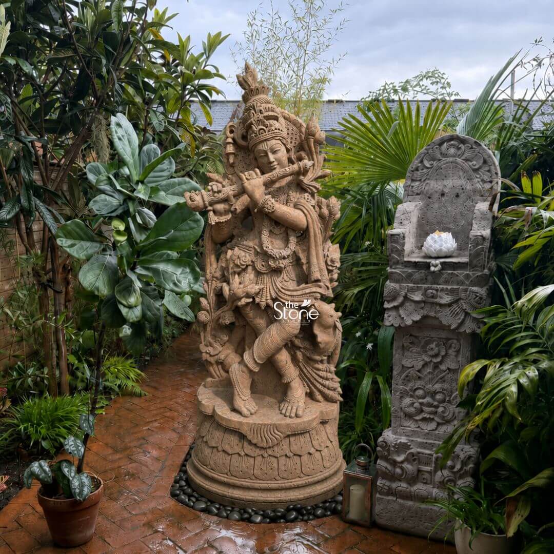 6 Feet Large Krishna Idol 3 A detailed medium shot captures a majestic six-foot-tall hand-carved statue of Lord Krishna sculpted from a warm reddish-brown sandstone positioned as a stunning focal point within a lush outdoor garden. The statue depicts Krishna in a graceful dancing tribhanga posture standing on an intricately carved circular lotus pedestal. He is playing a bamboo flute held to his lips with both hands conveying a sense of divine melody and peace. The sculpture exhibits exceptional artisan detailing with elaborate shallow-relief carvings of traditional jewelry a towering tiered crown garlands and flowing garments all meticulously rendered in the natural granular texture of the sandstone. The Krishna statue is situated at the end of a weathered brick pathway surrounded by a thriving array of tropical and temperate plants including large fiddle-leaf figs feathery palms and various ferns. This dense greenery creates a secluded verdant sanctuary that highlights the statue's sacred presence and demonstrates its natural integration into a sophisticated landscaping scheme. To the right of the Krishna figure stands a separate ornate Balinese-style stone plinth or shrine also hand-carved with detailed floral and faunal motifs. This complementary piece features a small white carved lotus flower ornament on its top surface further enhancing the spiritual and aesthetic atmosphere of the garden corner. The entire arrangement rests on a bed of small dark pebbles that define the statue's base and border the brick path. To the front right of the shrine plinth a traditional metal and glass lantern sits adding a warm inviting touch. The background shows more dense garden foliage and the edge of a house under an overcast sky providing a realistic domestic context for this elegant landscape feature. The natural diffused daylight illuminates the scene, casting soft defined shadows that emphasize the rich textures of the hand-carved sandstone and the complex forms of the surrounding plants making the statue a captivating element in the garden landscape.