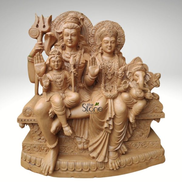 large, intricately carved stone sculpture depicts the revered Hindu divine family, known as the Shiva Parivar, seated on an elaborate throne. The artwork is rendered in a warm, light tan or sandalwood-colored stone, possibly sandstone or a similar natural material, highlighting its exquisite craftsmanship and traditional Indian aesthetic. From left to right, the central figure of Lord Shiva is depicted with a serene expression, his characteristic matted hair (jatamukuta) adorned with a crescent moon and a miniature figure of Ganga, and a coiled cobra resting on his left shoulder. He holds his iconic trident (trishula) in his raised upper left hand, while his lower right hand is raised in the Abhaya Mudra, a gesture of reassurance and blessing. Seated gracefully on Shiva's left thigh is Lord Kartikeya (also known as Skanda or Murugan), a youthful figure adorned with jewelry. He holds a long staff or vel in his left hand and also offers the Abhaya Mudra with his right hand. To Shiva's right, his consort, Goddess Parvati, is depicted with an elegant posture, wearing an ornate crown (kirita-mukuta) and beautiful, flowing garments. Her right hand is similarly raised in the Abhaya Mudra, symbolizing protection and peace. Nestled on Parvati's right thigh is their beloved son, Lord Ganesha, instantly recognizable by his elephant head with a gracefully curled trunk, a prominent pot belly, and holding a modak (sweet) in one hand. His right hand also extends in a gesture of blessing. The shared pedestal or throne on which the deities are seated is highly decorative, featuring intricate carvings including sun-like archways behind the heads of Shiva and Parvati, detailed scrollwork, floral motifs, and traditional Hindu iconography along the multi-tiered base, possibly including makara (mythical sea creatures) or lion faces. The overall composition conveys a sense of divine harmony, familial love, and spiritual grace, making it an ideal representation of sacred art for a temple, spiritual space, or home decor. The sculpture is presented against a plain, light grey background, which emphasizes the fine details and the natural beauty of the stone. A discreet watermark "the Stone Studio" is visible in the lower right, indicating the artisan or vendor.