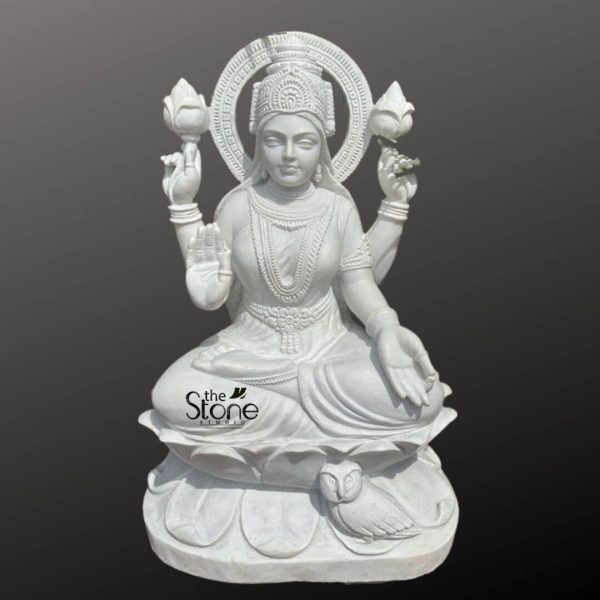 A majestic three-foot white marble statue of Maa Lakshmi, the Hindu goddess of wealth, prosperity, and fortune, is elegantly presented against a dark, contrasting background that highlights its pristine white finish. The goddess is depicted seated gracefully in a lotus posture (padmasana) upon a meticulously carved double-lotus pedestal. The entire sculpture radiates a serene and opulent aura. Maa Lakshmi is adorned with exquisitely sculpted traditional Indian jewelry, including multiple layered necklaces, intricate earrings, armlets, and bangles, all rendered in the same luminous white marble, showcasing the fine details of the carving. Her four arms are positioned with symbolic gestures and objects. Her lower right hand is extended in the gesture of blessing (varada mudra), often depicted with coins flowing from her palm, though not explicitly shown here. Her lower left hand rests gently on her lap, also in a relaxed gesture. Her upper two hands each hold a fully bloomed lotus flower, symbolizing purity, beauty, and spiritual enlightenment. A circular prabhavali (aura or halo) with delicate traditional patterns frames her head, emphasizing her divine presence. Maa Lakshmi's facial features are serene and benevolent, with finely sculpted eyes, nose, and lips that convey compassion and grace. Her hair is styled traditionally, topped with an ornate crown that signifies her divine royalty. Notably, a small, intricately carved owl, her traditional vahana (mount), is depicted seated at the base of the lotus pedestal on her right side, symbolizing wisdom and watchful discretion. The smooth, polished texture of the white marble, combined with the play of light and shadow, accentuates the statue's intricate details and its connection to the article's focus on a 3 feet Marble Maa Lakshmi Statue.