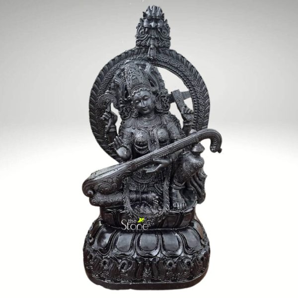 A majestic, five-foot-tall black stone statue of Maa Saraswati is depicted in a studio shot against a clean white background. The goddess is seated gracefully on a lotus pedestal, holding her Veena, a traditional Indian string instrument. Her four arms are adorned with intricate jewelry; two hands gently hold the Veena, while the upper right hand holds a rosary (mala) and the upper left hand holds a sacred manuscript. A large, ornate arch or Prabhavali frames her head, featuring detailed carvings that resemble flames or foliage, topped with a kirttimukha, a mythical monstrous face. The goddess's crown is elaborate, with delicate carvings, and her serene face features finely sculpted eyes, nose, and lips, conveying wisdom and tranquility. Her traditional attire, a sari, is sculpted with drapes and folds, enhancing the lifelike quality of the statue. The lotus base is intricately carved with layers of petals, and the overall craftsmanship showcases the fine details and textures of the black stone, highlighting the artistry and reverence associated with this depiction of Maa Saraswati. This image would be relevant for articles discussing Hindu deities, traditional Indian art, spiritual decor, or the symbolism of Saraswati Maa.