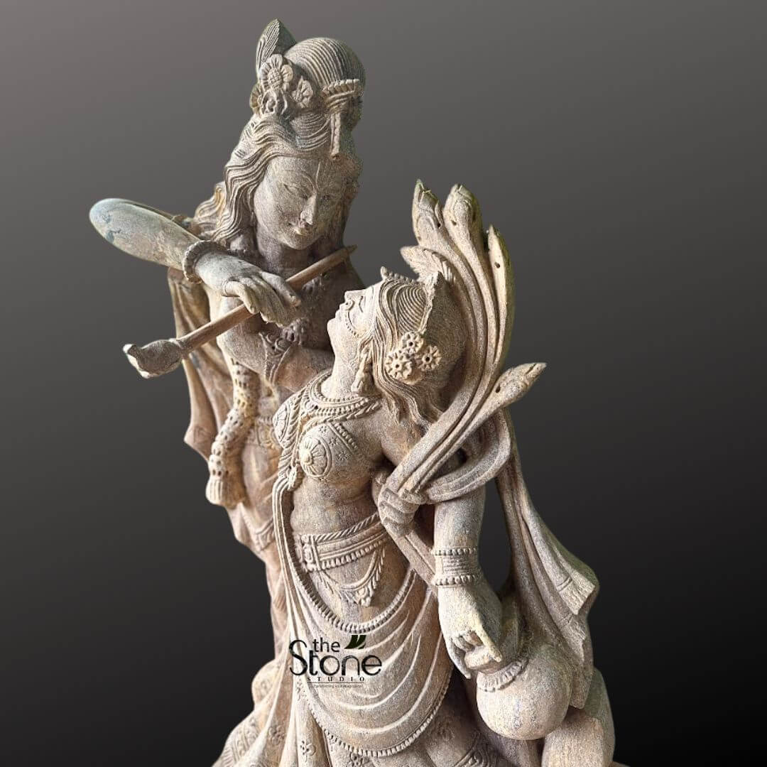Dancing Radha Krishna Stone Statue 3.5ft 2 A captivating close-up studio shot of a stone idol depicting the divine couple Radha and Krishna in a dynamic dancing embrace, set against a dark, contrasting grey background. The sculpture, crafted from a light-colored, possibly sandstone, showcases incredibly intricate detailing that emphasizes the romantic and devotional essence of their eternal dance. Lord Krishna, positioned slightly behind and above Radha, gazes tenderly down into her eyes while playing his iconic flute. The flute is held gracefully to his lips, and his posture suggests a melodic sway. Krishna is adorned with a detailed peacock feather in his crown, along with finely sculpted traditional jewelry, including necklaces, armlets, and bangles. His flowing attire, particularly his dhoti, is carved with expressive folds that convey movement and grace. Maa Radha is sculpted in front of Krishna, her body gracefully arched and tilted upwards, responding to his music and meeting his loving gaze. She is depicted in a pose that suggests elegant movement, with her traditional lehenga or skirt carved to show beautiful drapes and patterns, enhancing the sense of dance. Radha is sumptuously adorned with multiple necklaces, earrings, bangles, and an ornate crown or hairpiece, all meticulously detailed. Her left hand appears to be holding a small pot or musical instrument, further enhancing the dance theme. The texture of the stone is visible, highlighting the artisan's skill in rendering delicate features and flowing garments. The entire idol conveys profound divine love, harmony, and the joyous spirit of their celestial dance, perfectly aligning with the article's focus on a Dancing Radha Krishna Idol. The play of light and shadow on the textured stone further enhances the depth and emotional resonance of this sacred depiction.