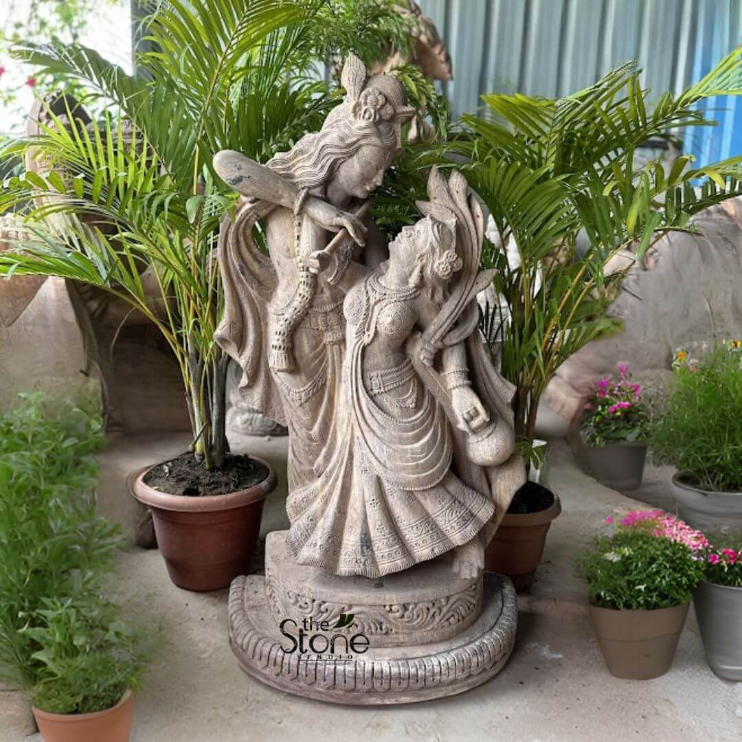 Dancing Radha Krishna Stone Statue 3.5ft 3 A beautifully crafted stone statue depicting the divine couple Radha and Krishna in a dynamic dancing pose, ideally suited for a garden setting, is presented amidst lush green potted plants. The statue, appearing to be made of light-colored, possibly sandstone, stands on a paved or earthy surface, surrounded by various tropical plants in terracotta pots, creating a serene and natural backdrop. Lord Krishna stands taller and slightly behind Radha, with his head tilted towards her, gazing affectionately into her eyes. He is playing his characteristic flute, held gracefully to his lips. Krishna is adorned with a detailed peacock feather in his crown, along with traditional jewelry like necklaces, armlets, and bangles, all finely sculpted into the stone. His dhoti (lower garment) flows dynamically, suggesting elegant movement and rhythm. Maa Radha is positioned in front of Krishna, her body gracefully arched and swaying, seemingly captivated by the music of his flute. Her head is tilted upwards, meeting Krishna's loving gaze with adoration and devotion. She is dressed in a traditional lehenga or skirt, which is also intricately carved with flowing folds and patterns that convey a sense of motion and fluidity. Radha is richly adorned with necklaces, earrings, bangles, and a crown or hair ornament, all meticulously detailed. Her left hand appears to hold a small pot or a subtle gesture related to dance. The base of the statue is a round, multi-tiered pedestal with carved floral and geometric patterns, providing a stable and aesthetic foundation. The entire sculpture conveys a profound sense of divine love, harmony, and eternal dance, perfectly capturing the essence of a Dancing Radha Krishna Statue for Garden. The natural lighting and lush surroundings enhance the spiritual and aesthetic appeal of the stone, making it a harmonious addition to an outdoor space