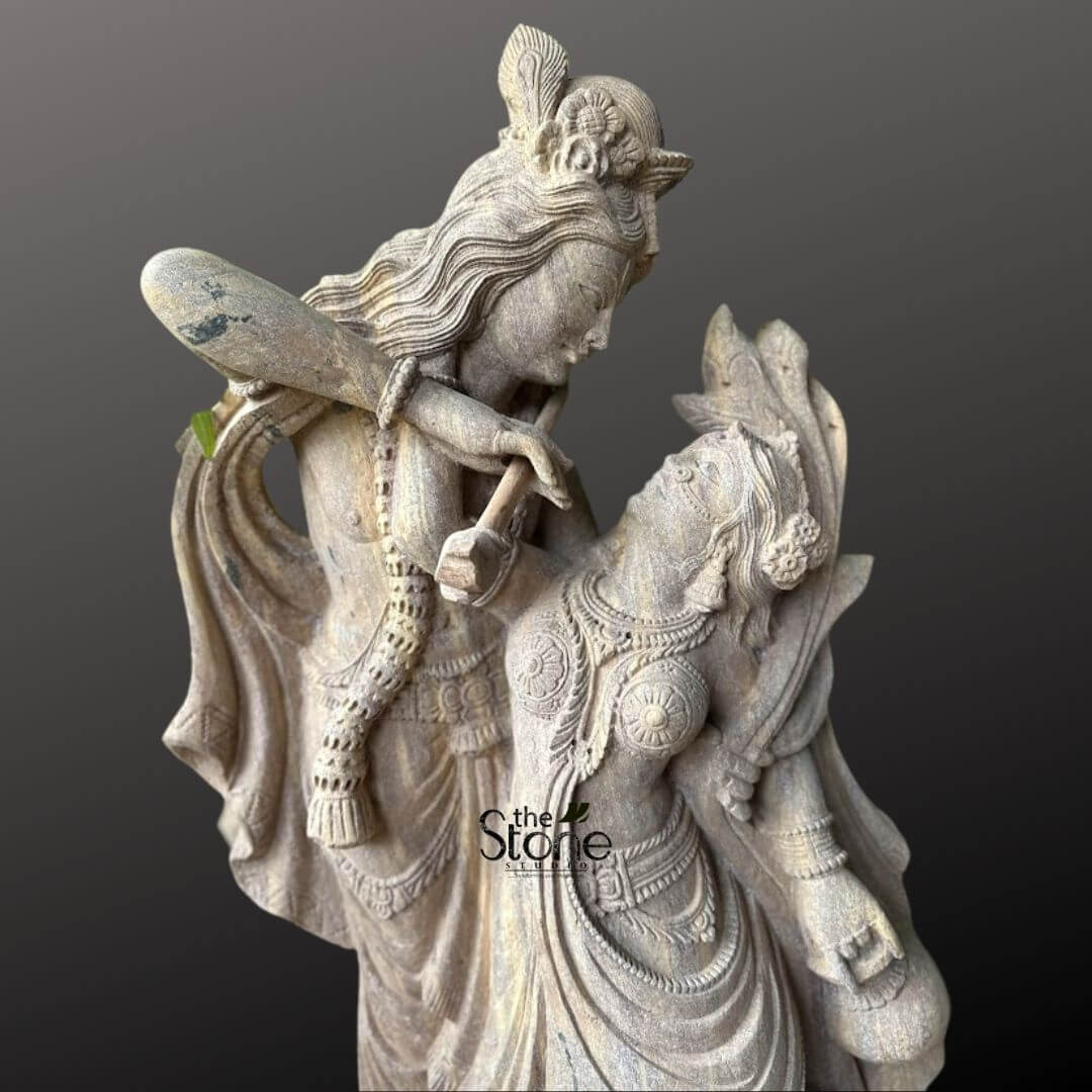 Dancing Radha Krishna Stone Statue 3.5ft 4 A captivating close-up studio shot of a stone statue depicting the divine couple Radha and Krishna in an intimate and dynamic dancing pose, set against a deep, contrasting grey background. The sculpture, crafted from a light-colored, textured stone, possibly sandstone, showcases exceptional artistry and intricate detailing that emphasizes the romantic and devotional essence of their eternal dance. Lord Krishna, positioned slightly behind and above Radha, gazes tenderly down into her eyes while playing his iconic flute. The flute is held gracefully to his lips, and his posture suggests a melodic sway, with his body slightly bent. Krishna is adorned with a detailed peacock feather in his crown, along with finely sculpted traditional jewelry, including necklaces, armlets, and bangles. His flowing attire, particularly his dhoti and sash, is carved with expressive folds that convey movement and grace. Maa Radha is sculpted in front of Krishna, her body gracefully arched and tilted upwards, responding to his music and meeting his loving gaze with profound adoration. She is depicted in a pose that suggests elegant movement, with her traditional lehenga or skirt carved to show beautiful drapes and intricate patterns, enhancing the sense of dance and fluidity. Radha is sumptuously adorned with multiple necklaces, prominent earrings, bangles, and an ornate crown or hairpiece, all meticulously detailed. Her left hand appears to be holding a small pot or a subtle musical instrument, further enriching the dance theme. The texture of the stone is highly visible, with subtle variations and natural imperfections that add to the sculpture's character and antique appeal. The artisan's skill in rendering delicate facial features, flowing garments, and intricate jewelry is evident. The entire idol conveys profound divine love, harmony, and the joyous spirit of their celestial dance, perfectly aligning with the article's focus on a Dancing Radha Krishna Statue. The interplay of light and shadow on the textured stone further enhances the depth and emotional resonance of this sacred depiction.
