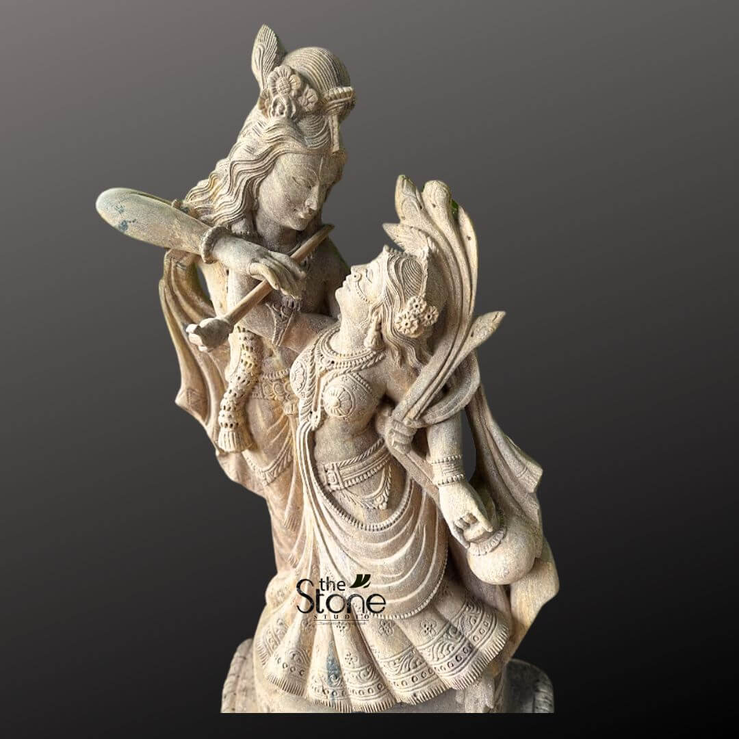 Dancing Radha Krishna Stone Statue 3.5ft 5 A captivating close-up studio shot of a stone idol depicting the divine couple Radha and Krishna in an intimate and dynamic dancing pose, set against a deep, contrasting grey background. The sculpture, crafted from a light-colored, textured stone, possibly sandstone, showcases exceptional artistry and intricate detailing that emphasizes the romantic and devotional essence of their eternal dance. Lord Krishna, positioned slightly behind and above Radha, gazes tenderly down into her eyes while playing his iconic flute. The flute is held gracefully to his lips, and his posture suggests a melodic sway, with his body slightly bent. Krishna is adorned with a detailed peacock feather in his crown, along with finely sculpted traditional jewelry, including necklaces, armlets, and bangles. His flowing attire, particularly his dhoti and sash, is carved with expressive folds that convey movement and grace. Maa Radha is sculpted in front of Krishna, her body gracefully arched and tilted upwards, responding to his music and meeting his loving gaze with profound adoration. She is depicted in a pose that suggests elegant movement, with her traditional lehenga or skirt carved to show beautiful drapes and intricate patterns, enhancing the sense of dance and fluidity. Radha is sumptuously adorned with multiple necklaces, prominent earrings, bangles, and an ornate crown or hairpiece, all meticulously detailed. Her left hand appears to be holding a small pot or a subtle musical instrument, further enriching the dance theme. The texture of the stone is highly visible, with subtle variations and natural imperfections that add to the sculpture's character and antique appeal. The artisan's skill in rendering delicate facial features, flowing garments, and intricate jewelry is evident. The entire idol conveys profound divine love, harmony, and the joyous spirit of their celestial dance, perfectly aligning with the article's focus on a Dancing Radha Krishna Stone Idol. The interplay of light and shadow on the textured stone further enhances the depth and emotional resonance of this sacred depiction.