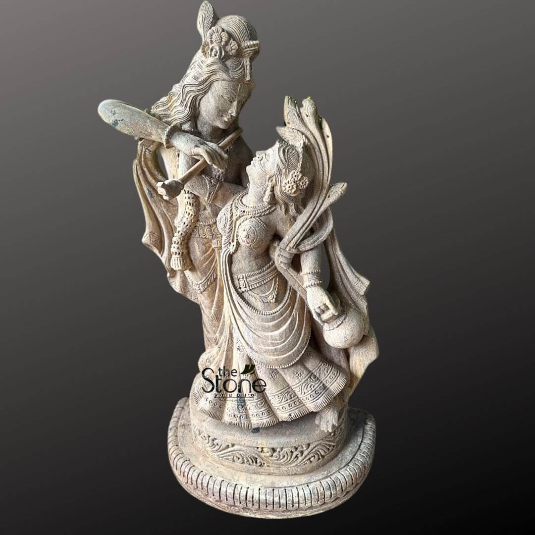 Dancing Radha Krishna Stone Statue 3.5ft 6 A beautifully carved stone sculpture of the divine couple Radha and Krishna, depicted in a dynamic and intimate dancing pose, is showcased against a deep, contrasting grey background. The sculpture, crafted from a light-colored, textured stone, possibly sandstone, exhibits intricate detailing that highlights the romantic and playful essence of their celestial dance. Lord Krishna stands taller and slightly behind Radha, with his head tilted towards her, gazing affectionately into her eyes. He is playing his characteristic flute, which is held gracefully to his lips. Krishna is adorned with a peacock feather in his crown, along with traditional jewelry like necklaces, armlets, and bangles, all finely sculpted into the stone. His dhoti (lower garment) flows dynamically, suggesting elegant movement. Maa Radha is positioned in front of Krishna, her body gracefully arched, seemingly swaying to the music of his flute. Her head is tilted upwards, meeting Krishna's gaze with adoration and devotion. She is dressed in a traditional lehenga or skirt, which is also sculpted with intricate folds and patterns to convey a sense of motion and fluidity. Radha is richly adorned with necklaces, earrings, bangles, and a crown or hair ornament, all meticulously detailed. Her left hand appears to be holding a small pot or a subtle musical instrument, further enriching the dance theme. The base of the sculpture is a round, multi-tiered pedestal with carved floral and geometric patterns, providing a stable and aesthetic foundation for the dynamic pose. The entire sculpture conveys a sense of profound divine love, harmony, and eternal dance, capturing the essence of the Dancing Radha Krishna Stone Sculpture with remarkable artistry and emotion. The texture of the stone, with its natural variations and subtle aging, adds to the sculpture's antique and sacred appeal.