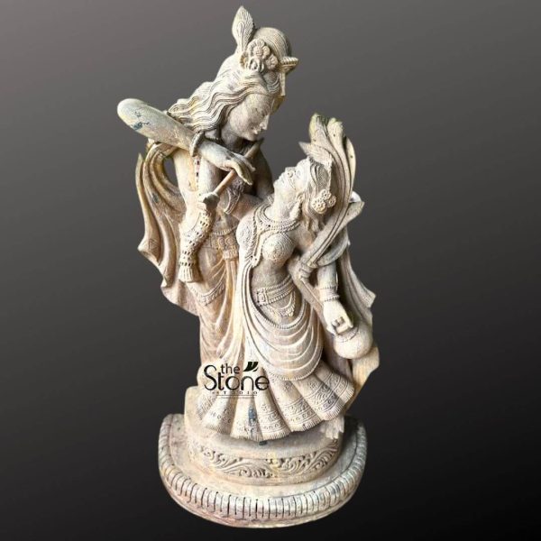 A beautifully carved stone statue of the divine couple Radha and Krishna, depicted in a dancing pose, is showcased against a deep, contrasting grey background. The statue, appearing to be crafted from a light-colored, possibly sandstone, exhibits intricate detailing that highlights the romantic and playful essence of their celestial dance. Lord Krishna stands taller and slightly behind Radha, with his head tilted towards her, gazing affectionately into her eyes. He is playing his characteristic flute, which is held gracefully to his lips. Krishna is adorned with a peacock feather in his crown, along with traditional jewelry like necklaces, armlets, and bangles. His dhoti (lower garment) flows dynamically, suggesting movement. Maa Radha is positioned in front of Krishna, her body gracefully arched, seemingly swaying to the music of his flute. Her head is tilted upwards, meeting Krishna's gaze with adoration. She is dressed in a traditional lehenga or skirt, which is also sculpted with intricate folds and patterns to convey a sense of motion and fluidity. Radha is richly adorned with necklaces, earrings, bangles, and a crown or hair ornament, all finely detailed. Her attire and jewelry are intricately carved, reflecting traditional Indian aesthetics. The base of the statue is a round, multi-tiered pedestal with carved patterns, providing a stable foundation for the dynamic pose. The entire sculpture conveys a sense of divine love, harmony, and eternal dance, capturing the essence of the Dancing Radha Krishna Stone Statue with remarkable artistry and emotion. The texture of the stone, with its natural variations and subtle aging, adds to the statue's antique and sacred appeal.