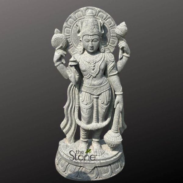 A magnificent 4-foot tall grey stone statue of Lord Vishnu, depicted standing elegantly and holding his traditional attributes. The statue showcases Vishnu with four arms, symbolizing his omnipresence and power. In his upper right hand, he holds the Sudarshana Chakra (discus), and in his upper left hand, he holds the Panchajanya Shankha (conch shell). His lower right hand holds the Kaumodaki Gada (mace), and his lower left hand is positioned in a gesture of blessing or holding a lotus, though the specific detail might be subtly carved. Lord Vishnu is adorned with an elaborate crown (Kiritamukuta), intricate necklaces, armlets, wristlets, and anklets, all meticulously carved into the stone. He wears a beautifully draped dhoti and a sacred thread (Yajnopavita). A prominent halo (Prabhāmaṇḍala) with intricate patterns is carved behind his head, signifying his divinity. The statue stands on a multi-layered lotus pedestal, adding to its sacred aesthetic. The carving is highly detailed, highlighting the serene expression on Vishnu's face, the textures of his clothing, and the symbolic attributes. The background is a solid dark grey, making the lighter grey stone statue stand out as the central focus.
