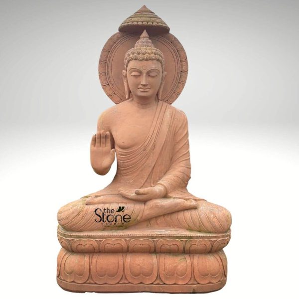 A majestic 7-foot stone Buddha statue, crafted from a warm reddish-brown natural stone, is presented in a studio shot against a clean, light gray background, highlighting its grand presence and intricate details. The Buddha is depicted in a meditative seated posture, with legs crossed in Padmasana, resting gracefully on a double lotus pedestal. His right hand is raised in the Abhaya Mudra (gesture of fearlessness and reassurance), while his left hand rests in his lap, palm upward, in a gesture of meditation. His face exudes serene contemplation, with downcast eyes, a gentle smile, and finely carved features that convey profound peace. The Buddha's head is adorned with tightly coiled hair, leading up to an Ushnisha, a protuberance symbolizing wisdom. Behind his head, a large circular halo or Prabhavali features a subtle, radiating pattern, symbolizing his enlightened state. Above the halo, a multi-tiered parasol-like structure provides an elegant canopy. The flowing drapery of his monastic robe is sculpted with natural folds, accentuating the contours of his body. The lotus base is intricately carved with layers of petals, grounding the impressive figure. The overall craftsmanship and natural stone finish make this a striking and reverent representation, ideal as a centerpiece for a large garden, temple, or spacious interior.