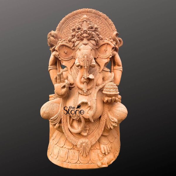 A grand and imposing large Ganesha statue, carved from reddish-brown sandstone, is showcased against a deep, solid grey background, highlighting its suitability as a prominent outdoor feature. Lord Ganesha, the revered Hindu deity of wisdom, prosperity, and remover of obstacles, is depicted in a traditional seated posture, with one leg folded and the other slightly raised, conveying both majesty and approachability. The statue features Ganesha with his iconic elephant head, adorned with an elaborate, multi-tiered crown that includes intricate traditional motifs and a central design. His large, fan-like ears are prominently sculpted, and his trunk is elegantly curled towards his left, reaching for a modak (sweet dumpling) held in one of his hands. His eyes are carved with a serene and benevolent expression, conveying wisdom and calm. Ganesha is portrayed with four arms, each holding symbolic attributes. His lower right hand is raised in the abhaya mudra, a gesture of blessing and reassurance, with finely detailed palm lines. His lower left hand gently holds a bowl of modaks, symbolizing abundance and the fruits of spiritual labor. His upper right hand grasps an ankusha (goad), representing his ability to guide devotees on the righteous path, while his upper left hand holds a pasha (noose), symbolizing control over worldly attachments. He is richly adorned with sculpted jewelry, including a prominent necklace, armlets, and anklets, all intricately carved to mimic traditional designs. A sacred thread (yagnopavitam) drapes across his chest, and his robust, pot-bellied form, symbolizing prosperity, is artfully rendered. A large, ornate prabhavali (aura or halo) with detailed carvings frames his head, enhancing his divine presence. The entire sculpture is rich in texture from the sandstone, with visible chisel marks adding to its artisanal charm and emphasizing its robust nature for outdoor placement, aligning with the Big Ganesha Statue For Outdoors theme.