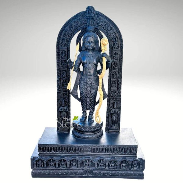 A magnificent black stone statue of Ram Lalla (Lord Rama in his child form) stands elegantly within an intricately carved archway, presented in a studio shot against a clean white background. The central figure of Ram Lalla is sculpted with a youthful yet divine presence, holding a golden bow in his left hand and a golden arrow in his right, poised for dharma. He is adorned with delicate jewelry, including necklaces, armlets, and anklets, and wears a traditional dhoti with finely sculpted pleats and patterns. His serene facial features, including large eyes and a gentle smile, convey both innocence and divine power. His hair is styled in a traditional manner, and a subtle halo or divine aura surrounds his head. The statue stands on a lotus pedestal, which is part of a larger, multi-tiered black stone base. This base features intricate carvings, including a row of smaller deities or devotees along its lower edge. The elaborate archway (Prabhavali) that frames Ram Lalla is a masterpiece of carving, featuring detailed patterns, possibly depicting celestial beings, floral motifs, and architectural elements. The arch is supported by two ornate pillars with figures carved at their bases. The entire sculpture, from the base to the arch, is crafted from dark, lustrous black stone, highlighting the intricate details and the reverence associated with this depiction of Lord Rama. The golden accents on the bow and arrow provide a striking contrast against the dark stone, emphasizing their significance. This statue beautifully captures the essence of Ram Lalla, combining youthful grace with divine majesty.