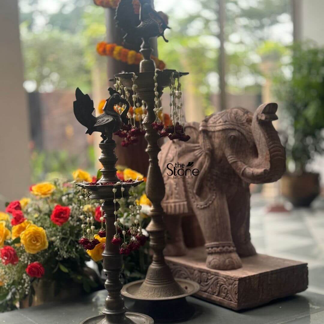 A beautifully arranged tableau showcases traditional Indian decor, featuring a 2-foot elephant statue as a prominent element, indicative of Elephant Statue placement in Vijayawada. The scene is set indoors, with natural light filtering through large windows in the background, revealing lush greenery and perhaps a building exterior. In the foreground, two elegant brass oil lamps (diyas) stand tall and adorned. The left lamp features a peacock motif at its apex, a traditional Indian design, and is decorated with delicate strings of white flowers and small red beads. The right lamp is a multi-tiered traditional brass lamp, also embellished with similar floral strings. Both lamps are placed on a dark, possibly marble or granite, surface. Behind and slightly to the right of the lamps, a magnificent 2-foot elephant statue, carved from reddish-brown sandstone, stands majestically on a rectangular base. The elephant is intricately detailed, depicting fine carvings of traditional caparison (decorative coverings) on its back and sides, with its trunk raised in a welcoming or auspicious gesture. The texture of the sandstone is visible, giving the statue a rustic yet refined appearance. To the left of the elephant, a vibrant bouquet of fresh roses in shades of red and yellow adds a splash of color and freshness to the arrangement. In the blurred background, through the windows, more green plants and natural light are visible, suggesting a serene and elegant interior space where traditional elements are celebrated. The overall composition highlights the cultural significance and aesthetic appeal of the elephant statue within a refined Indian home or commercial setting.
