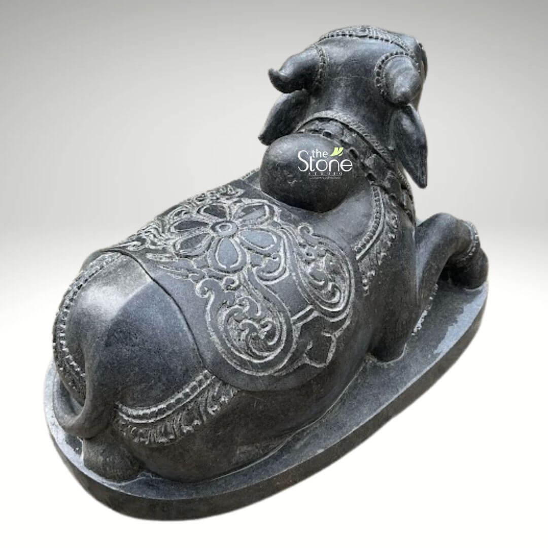 2ft Nandi Bull Stone Idol 4 A rear-view, slightly elevated shot captures a 2-foot black stone Nandi statue set against a plain light-colored background. The statue depicts the sacred bull Nandi in a reclining position traditional for its depiction as Shiva's mount. The black stone material gives it a solemn and earthy appearance. The Nandi is richly adorned with intricate carvings across its back and sides. A large circular floral pattern is prominent on its back surrounded by swirling ornate designs that extend towards its haunches and a decorative saddle-like covering. Smaller patterns also embellish its neck and head visible from this angle. The texture of the stone is smooth yet allows the detailed carvings to stand out. The statue rests on a simple oval-shaped base also made of the same dark material. This detailed image emphasizes the traditional craftsmanship and spiritual significance of the 2-foot black stone Nandi statue a key subject of the accompanying article.