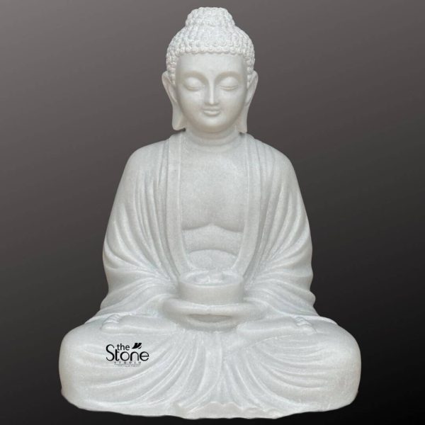 A serene 2-foot white marble Buddha statue depicted in a seated meditation pose against a dark background. The statue is meticulously hand-carved from high-quality marble, featuring a smooth, luminous white finish. The Buddha’s face reflects deep tranquility with closed eyes and a gentle, peaceful expression, crowned by traditional snail-shell curls and an ushnisha. He is shown holding a ceremonial alms bowl in both hands resting on his lap, a classic symbol of spiritual nourishment. The monastic robes are draped gracefully with realistic, flowing folds that highlight the fine craftsmanship of the stone carving. This elegant 2-foot statue is a perfect addition for those looking to bring serenity and spiritual harmony to their home, meditation room, or garden landscape.