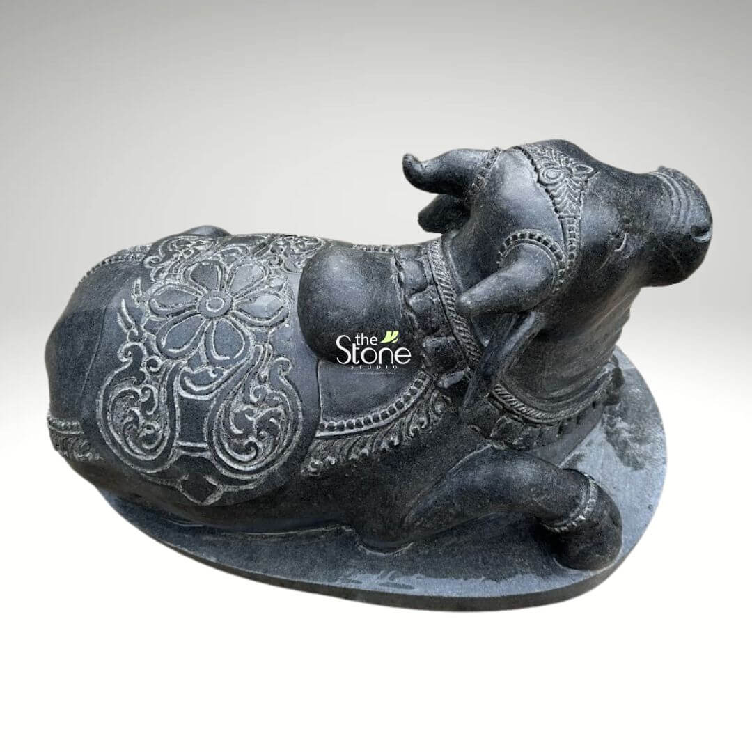 2ft Nandi Bull Stone Idol 6 An exquisitely hand-carved 2-foot black stone Nandi bull sculpture masterfully designed as a sacred centerpiece for a home pooja room or temple sanctuary. This top-down rear perspective highlights the exceptional 360-degree craftsmanship of the piece focusing on the intricate decorative blanket or caparison draped over the bull’s back. The sculpture crafted from premium natural black stone features a large detailed floral mandala at its center surrounded by rhythmic geometric and scrollwork patterns etched in relief. Lord Nandi the loyal vehicle of Lord Shiva, is depicted in a stable and serene recumbent pose with visible ornamental bands around his neck and an ornate headpiece between his horns. The deep charcoal-black finish of the stone provides a sophisticated rustic aesthetic that highlights the heritage of traditional Indian stone masonry. Resting on a solid integrated oval pedestal, this 2ft Nandi idol is a durable and auspicious addition to any spiritual space designed to symbolize devotion strength and the invitation of positive energy into the home.