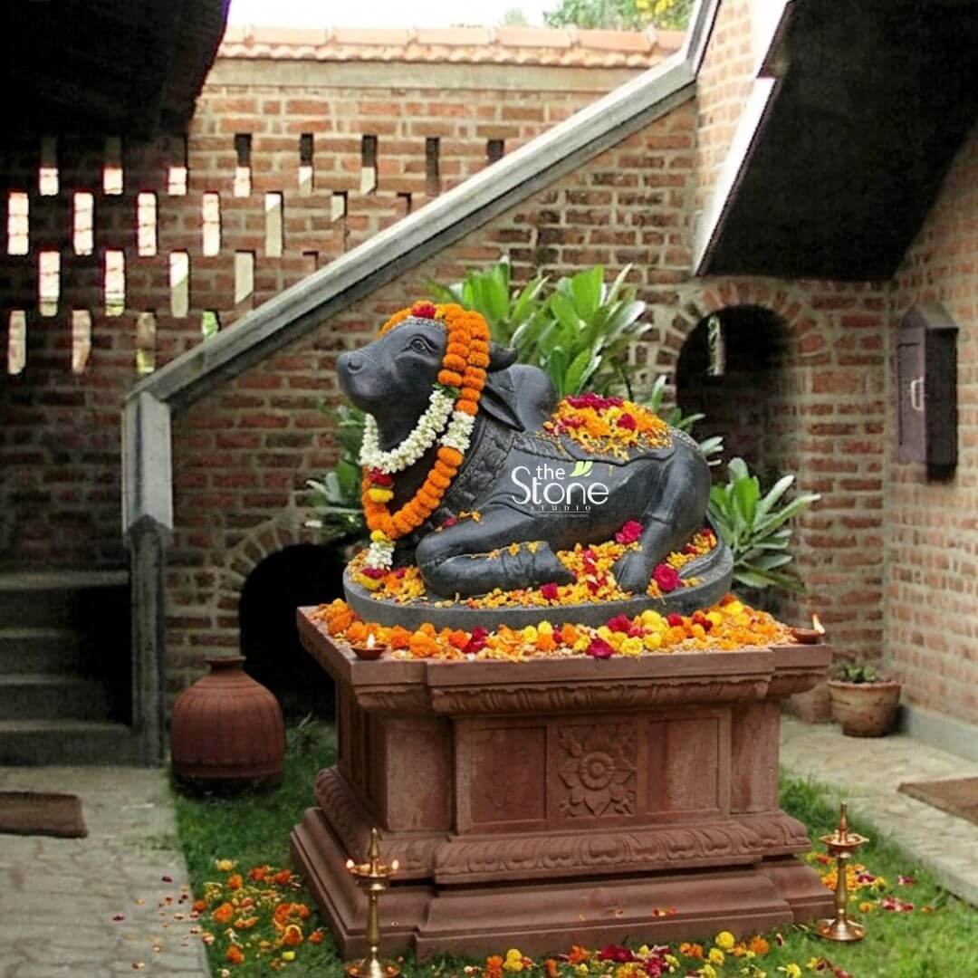 2ft Nandi Bull Stone Idol 3 An exquisitely hand-carved 2-foot tall black stone Nandi Maharaj sculpture masterfully crafted from premium natural stone to serve as a majestic spiritual centerpiece. The sacred bull known as the loyal vehicle of Lord Shiva is depicted in a serene and stable recumbent posture atop an intricately detailed multi-tiered reddish-brown pedestal featuring traditional relief carvings. The black stone Nandi idol is festively adorned for worship with thick, vibrant garlands of orange marigolds and delicate white jasmine blossoms. Loose red rose and yellow marigold petals are gracefully scattered across the deity’s back and around the circular stone base. The sculpture is artfully placed in an outdoor garden or courtyard setting set against a backdrop of a rustic brick wall and a contemporary staircase with unique geometric cut-outs. Flanking the base on the lush green grass are small polished brass oil lamps (diyas) creating a peaceful and auspicious atmosphere. This 2ft Nandi stone statue perfectly illustrates how heritage-style stone craftsmanship can be integrated into a modern home mandir or landscape design to invite strength, devotion and divine protection into the surroundings.