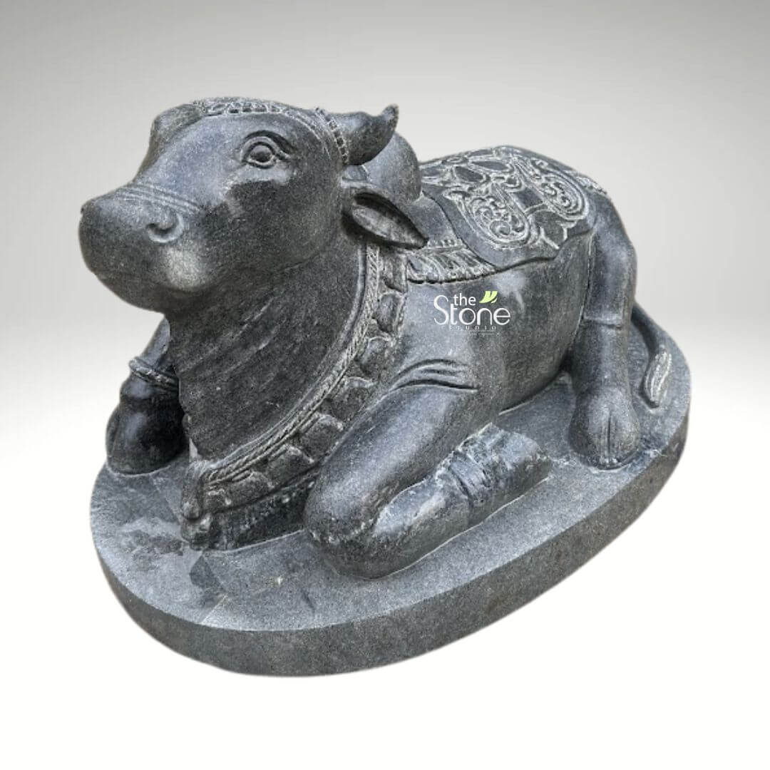 2ft Nandi Bull Stone Idol 5 An exquisitely hand-carved 2-foot tall black stone Nandi murti masterfully designed to serve as a majestic spiritual centerpiece for a temple entrance or home mandir. This black stone Nandi statue captures the sacred bull, the loyal vehicle (vahana) of Lord Shiva, in a serene and stable recumbent posture atop a solid, integrated oval pedestal. The sculpture showcases exceptional craftsmanship with highly intricate detailing. Nandi is adorned with traditional temple ornaments including multiple layers of carved bell necklaces around its sturdy neck and a decorative headpiece positioned between its ears. A richly detailed caparison (decorative blanket) featuring rhythmic floral and geometric relief patterns is draped across its back and prominent hump, reflecting the grandeur of heritage Indian stone masonry. The deep charcoal hue and naturally grainy texture of the stone emphasize the artisan’s attention to detail, giving the idol a timeless, rustic appeal. This durable and weather-resistant 2ft Nandi sculpture is ideal for creating an atmosphere of devotion and strength in any sacred space, whether positioned at the threshold of a grand temple or within a personal prayer room, designed to invite divine protection and peace into the environment.