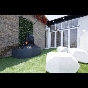 A majestic matte black Adiyogi statue, representing Lord Shiva with a white crescent moon, is the centerpiece of a contemporary rooftop garden in Pune, Maharashtra. The bust is thoughtfully placed within a curved, dark-textured water basin where a rainfall water feature cascades over it. The setup is set against a rustic brown stone wall featuring a vertical strip of lush green foliage with delicate white flowers directly behind the statue. Vibrant red blossoms drape from the top of the wall, adding a touch of color to the earthy tones. The ground is covered in bright green artificial turf, accented by large, white geometric seating designed to look like faceted pebbles. This serene outdoor arrangement exemplifies a modern spiritual retreat in an urban Maharashtra residence, blending traditional iconography with minimalist landscape design.