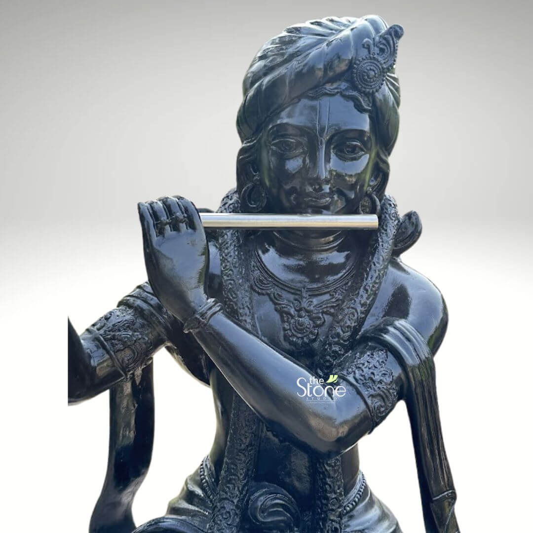 Lord Krishna with Cow Fibre Statue 4ft 6 A detailed upper-body view of an exquisitely crafted Bal Krishna fibre statue, showcasing a high-gloss black finish with a radiant, polished luster. The Hindu deity is depicted with a serene facial expression, gracefully holding a silver-toned flute to his lips in a meditative playing posture. Lord Krishna is adorned with highly detailed traditional ornaments, including an elaborate turban with a central crest, layered necklaces, and armlets featuring fine relief patterns. The reflective surface of the black fibre sculpture highlights the intricate craftsmanship of the attire and jewelry. This durable and lightweight spiritual icon beautifully captures the divine essence of the cowherd god, serving as an auspicious and artistic centerpiece for home temple decor or garden landscapes, often paired with a sacred cow to complete the traditional devotional scene.