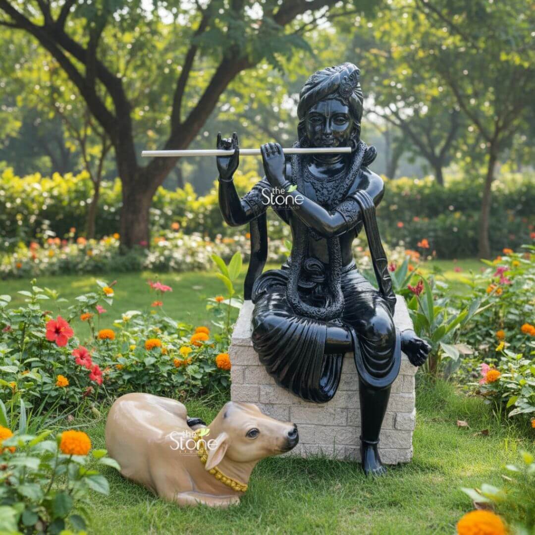Lord Krishna with Cow Fibre Statue 4ft 3 A serene and auspicious scene featuring a high-gloss black finish Krishna cow fibre murti as a spiritual centerpiece in a lush outdoor garden. The Lord Krishna statue is depicted in a graceful seated posture on a light-colored, textured stone pedestal, delicately holding a silver-toned flute to his lips. He is adorned with a detailed turban and traditional Indian jewelry, with the polished black surface reflecting the natural sunlight. Resting peacefully on the green grass at the base of the pedestal is a lifelike, light-brown calf adorned with a vibrant yellow beaded garland, symbolizing the divine bond between Krishna and the sacred cow. The entire setting is framed by a beautiful landscape of mature trees and blooming flowers, including bright orange marigolds and red hibiscus. This durable and weather-resistant fibre statue perfectly illustrates how a spiritual icon can be integrated into high-end garden decor to create a tranquil space for meditation and peace in a home sanctuary.
