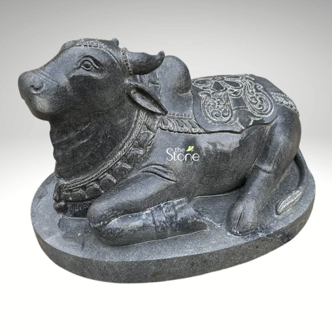 2ft Nandi Bull Stone Idol 7 An exquisitely hand-carved classic 2ft Nandi statue for home masterfully sculpted from premium natural black stone to serve as a majestic spiritual centerpiece. This 2-foot tall black stone Nandi idol depicts the sacred bull the loyal vehicle (vahana) of Lord Shiva in a serene and stable recumbent posture atop a solid, integrated oval pedestal. The sculpture showcases exceptional craftsmanship with highly intricate detailing. Nandi is adorned with traditional temple ornaments, including multiple layers of carved bell necklaces and beaded garlands around its sturdy neck. A richly detailed caparison featuring rhythmic floral relief patterns and fine borders is draped across its back and prominent hump, reflecting the grandeur of heritage Indian stone masonry. The deep charcoal-black hue and the naturally grainy texture of the stone emphasize the artisan's attention to detail, giving the idol a timeless, rustic appeal. This durable and weather-resistant 2ft Nandi sculpture is ideal for creating an atmosphere of devotion and strength in a home mandir, meditation room, or at the entrance of a house, designed to invite divine protection and peace into the environment.