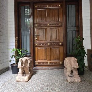 A pair of hand-carved stone elephant statues is placed symmetrically on either side of a grand home entrance, exemplifying a traditional and auspicious placement in Karnataka. The statues, crafted from textured light-brown natural stone, feature intricate carvings on their bodies and are positioned on solid rectangular bases. Each elephant is depicted with its trunk held in a natural, downward-curving pose, facing forward to greet visitors. The pair sits atop a grey patterned tiled porch floor, flanking a large, ornate dark wood door adorned with decorative brass studs and hardware. The background features white brick-textured walls and large windows with black metal security bars. Two lush green potted plants are tucked behind the statues on either side, adding a vibrant natural element to the entryway. This symmetrical arrangement of an elephant pair statue is a popular design choice for residences in Karnataka, believed to bring strength, protection, and positive energy to the household.