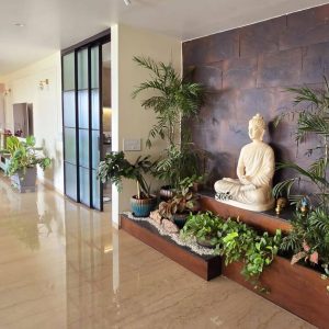 A tranquil interior space features a light-colored fiber Buddha statue, approximately three feet in height, seated in a meditative posture, serving as a peaceful focal point. The statue is elegantly positioned against a textured accent wall of dark, rectangular stone tiles. A variety of lush green plants, including tall palms and trailing foliage, are meticulously arranged in a long, dark wooden planter box that spans the length of the wall, creating an indoor garden effect. The polished, light-colored floor reflects the natural light, contributing to the bright and airy feel of the room. To the left, a modern black-framed glass sliding door leads to another area, while additional potted plants are visible further down the hallway, enhancing the serene and natural aesthetic. This setup highlights how a fiber Buddha statue can create a calm and stylish environment in a contemporary home in Gujarat.