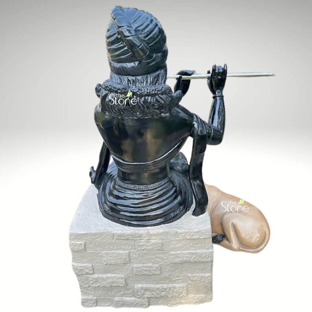 Lord Krishna with Cow Fibre Statue 4ft 7 A rear view of an intricately designed fibre statue of flute playing Krishna with a cow, showcasing the exceptional 360-degree craftsmanship of the sculpture. Lord Krishna is depicted in a seated posture atop a light grey pedestal featuring a realistic brick-stone texture. The deity is finished in a high-gloss black tone that highlights the detailed carvings of his traditional turban, wavy hair, and the fluid folds of his draped garments. Capturing the iconic musical gesture of the divine cowherd, Krishna’s hands are positioned as if playing a silver-toned flute. Resting at the base of the pedestal is a lifelike light-brown cow, adorned with a vibrant yellow beaded garland, symbolizing the divine protection and love for animals. This durable and lightweight fiber Krishna sculpture is a perfect spiritual centerpiece for contemporary home interiors, mandirs, or outdoor garden decor, offering a serene and auspicious presence from every perspective.