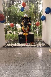 A stunning black Ganesh idol serves as a focal point in a celebratory indoor setting in Kolkata. The statue is gracefully draped in traditional floral garlands of yellow, white, and pink flowers. It sits on a dark pedestal surrounded by a carpet of scattered white jasmine petals on a polished stone floor. The festive atmosphere is enhanced by colorful blue, orange, and white balloons floating around the deity. A small, intricately hand-painted decorative pillar and a brass bowl filled with traditional laddoos are placed in front as offerings. In the background, large floor-to-ceiling glass panels offer a serene view of a lush tropical garden, beautifully blending the indoor celebration with Kolkata's natural greenery.