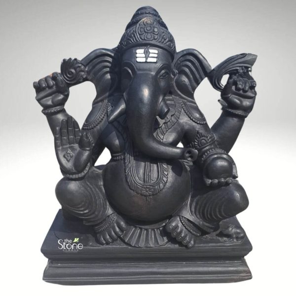 A masterfully hand-carved Ganesha idol for temple placement, crafted from smooth matte black stone, stands as a majestic spiritual centerpiece. The four-armed elephant-headed deity is depicted in a traditional seated posture atop a tiered rectangular pedestal. The sculpture showcases exquisite craftsmanship, featuring an intricately detailed crown, ornate necklaces, and fine bangles. One of his hands is raised in the Abhaya Mudra, a sacred gesture of protection and blessing, while another holds a bowl of sweet laddus. A prominent white tripundra mark is carved onto his forehead, adding to his divine appearance. With a calm, benevolent expression and a gracefully curved trunk, this black stone Ganesha idol is designed to bring a sense of peace and auspiciousness to any home mandir or temple sanctum.