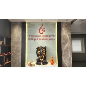 A majestic black stone Ganesha idol, reminiscent of the traditional spiritual artistry found in Deogarh, is elegantly placed at the center of a contemporary home temple. The idol, adorned with a vibrant yellow and orange flower garland, sits on a clean white platform. Behind the Ganesha statue, a large, softly backlit panel serves as a focal point, featuring a red Om symbol at the top and the sacred Ganesha shloka, Vakratunda Mahakaya, inscribed in Sanskrit. The serene prayer area is framed by dark vertical panels and flanked by polished grey marble walls with white veining, illustrating a modern puja unit design. To the right of the main deity sits a smaller idol of Shirdi Sai Baba dressed in orange, while traditional puja essentials, including a conch shell, are neatly arranged on the left. The warm, recessed ceiling lights and the glowing back panel create a tranquil and spiritually uplifting atmosphere, showcasing how a traditional Ganesha idol can be integrated into a sophisticated modern interior.
