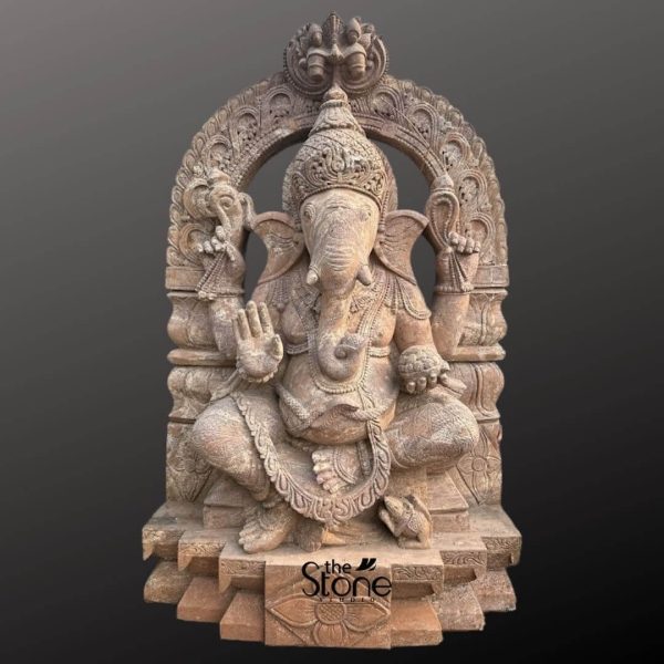 This magnificent 5-foot tall Lord Ganesha sculpture is an exquisite choice for a Ganesha Sculpture for Garden, hand-carved from premium brown sandstone to create a serene and auspicious outdoor sanctuary. The statue features a rich, earthy texture and detailed craftsmanship that brings out the timeless beauty of natural stone, making it a durable and weather-resistant centerpiece for any landscaping project. The deity is depicted in a traditional seated pose on a multi-tiered pedestal, conveying a sense of stability and calm. Lord Ganesha’s four arms hold significant spiritual symbols: his upper hands hold a rope and an axe to help devotees overcome ignorance and obstacles, while his lower left hand holds a modak, a traditional sweet representing the rewards of a spiritual life. His lower right hand is raised in the Abhaya Mudra, the gesture of fearlessness and blessing, offering protection and peace to all who enter the garden. Intricate carvings adorn the entire sculpture, from the elaborate, tiered crown on his elephant head to the floral patterns on the majestic Prabhavali (ornate arch) that frames him. At the base of the statue, his loyal vehicle, the mouse (Mooshika), is delicately carved, symbolizing humility and the ability to master one’s desires. This sandstone Ganesha statue is not only a profound spiritual icon but also a striking piece of garden art that will age gracefully, developing a unique patina over time while standing firm against the elements. It is an ideal focal point for a meditation corner, home entrance, or a lush green backyard, inviting positive energy and prosperity into the surroundings.