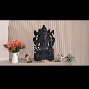 An exquisitely handcrafted 2-foot tall black stone Ganesha statue serves as a serene spiritual centerpiece in a modern Ahmedabad residence. The sculpture is carved from premium natural black stone with a refined matte finish, featuring intricate details on the deity's multi-tiered crown mukut, large expressive ears, and gracefully curved trunk. Lord Ganesha is depicted in a seated meditative pose on a decorative lotus base, with his right hand raised in the Abhaya Mudra, the universal gesture of protection and fearlessness. The blackstone Ganesha idol is artfully placed on a sleek, grey marble mantle against a contemporary beige fluted wall backdrop. It is elegantly framed by a white ceramic vase of vibrant orange lilies on one side and a delicate green potted plant on the other. Two miniature white and pink elephant figurines flank the main idol, adding a traditional touch to the minimalist setting. This devotional setup perfectly highlights how a traditional hand-carved Ganesha sculpture can seamlessly integrate into high-end urban home interiors, bringing peace, prosperity, and an aura of divine stability to the living space.