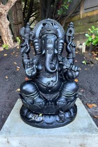 A majestic Ganesha statue, meticulously hand-carved with a smooth charcoal-black finish, stands as a spiritual centerpiece in a serene garden in Canada. The elephant-headed deity is depicted in a traditional seated pose upon an ornate, multi-petaled lotus throne. His four arms hold sacred symbolic objects, with one palm held up in a blessing gesture that features a finely engraved 'Om' symbol. The statue showcases master craftsmanship through its elaborate crown, intricate necklaces, and a sacred cobra wrapped gracefully around the deity's belly. At the base of the lotus, Ganesha's divine vehicle, the small mouse (Mooshika), is depicted in a humble posture. Placed on a solid concrete plinth amidst dark garden mulch and surrounded by natural trees, this stone.