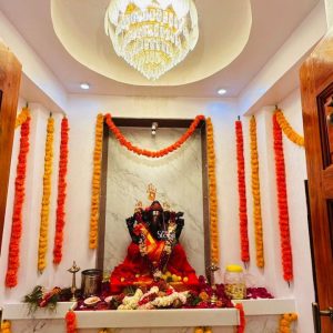 A divine black Ganesha statue is enshrined in a brilliantly lit temple sanctum in Sambhaji Nagar, Maharashtra. The idol is elegantly draped in a vibrant red silk vastra (cloth) and adorned with multiple garlands of fresh yellow and white blossoms. Set against a white marble-patterned backdrop featuring a sacred red handprint, the deity is the focal point of this spiritual space. The surrounding walls are festively decorated with long vertical and horizontal strings of orange and yellow marigold garlands, a traditional touch for temple celebrations in Maharashtra. At the base of the Ganesha idol, a carpet of pink rose petals is scattered among various offerings, including coconuts and floral arrangements. Two classic brass oil lamps flank the idol, while a grand crystal chandelier hangs from a recessed circular ceiling, casting a warm and opulent glow over the entire shrine.