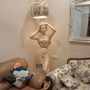A 5.5-foot fibre statue of a graceful lady, often used as a graceful angel statue for a home entrance, is beautifully positioned in a well-lit corner of a living room. Standing tall between a modern brown leather armchair and a classical floral-print sofa, the statue is illuminated by a warm, overhead light from a decorative, hand-painted pendant lamp. The figure is depicted in a timeless classical pose, adjusting her hair with both hands, while wearing a floor-length, flowing gown with masterfully sculpted drapery. The statue's light cream finish adds a touch of elegance to the neutral-toned room. To the side, a rustic white-framed mirror reflects the sophisticated atmosphere of this residential space, demonstrating how a large-scale fibre statue can transform a home foyer or entrance into a grand spiritual and artistic sanctuary.