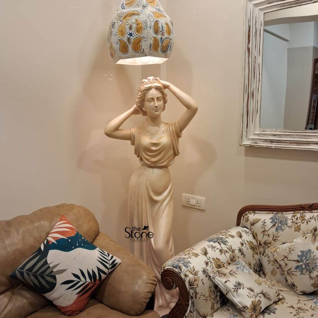 Angel Lady Statue for Resorts 5.5 Feet 4 A 5.5-foot fibre statue of a graceful lady, often used as a graceful angel statue for a home entrance, is beautifully positioned in a well-lit corner of a living room. Standing tall between a modern brown leather armchair and a classical floral-print sofa, the statue is illuminated by a warm, overhead light from a decorative, hand-painted pendant lamp. The figure is depicted in a timeless classical pose, adjusting her hair with both hands, while wearing a floor-length, flowing gown with masterfully sculpted drapery. The statue's light cream finish adds a touch of elegance to the neutral-toned room. To the side, a rustic white-framed mirror reflects the sophisticated atmosphere of this residential space, demonstrating how a large-scale fibre statue can transform a home foyer or entrance into a grand spiritual and artistic sanctuary.