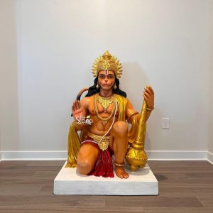 A vibrant and life-sized Lord Hanuman statue serves as a spiritual centerpiece in a modern indoor setting in Georgia. The Hindu deity is depicted in a powerful kneeling pose on a solid white square pedestal, set against a backdrop of minimalist light grey walls and contemporary dark wood flooring. The statue is richly colored with warm orange skin tones and features a serene yet strong expression. He wears a brilliant, sun-like golden crown and is adorned with intricate traditional jewelry, including tiered necklaces, armlets, and a white sacred thread. Hanuman's right hand is raised in the Abhaya Mudra, a universal gesture of protection and fearlessness, while his left hand firmly holds a large golden mace, or Gada, resting on the floor. He is draped in a bright yellow sash and a deep red loincloth, with his iconic long tail curving gracefully behind his head. This magnificent sculpture beautifully illustrates the integration of traditional Indian spiritual art into a contemporary Western home or community space in Georgia, creating a dedicated area for mindfulness and devotion.