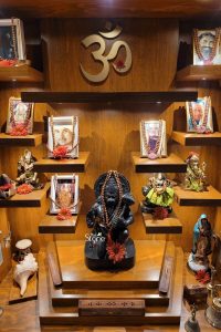 A beautifully crafted black stone Lord Hanuman statue serves as the spiritual centerpiece of an elaborate, multi-tiered dark wood home altar in Mexico. The deity is depicted in a devotional kneeling pose, holding his signature Gada (mace) and draped with a sacred Rudraksha mala. The entire space is bathed in warm, soft lighting from recessed spotlights, highlighting the rich textures of the wood and the diverse collection of religious icons. Surrounding the Hanuman statue are several floating shelves holding other Hindu deities, such as Lord Ganesha and Lord Shiva, along with framed portraits of spiritual gurus adorned with floral garlands. At the top center, a large golden 'Om' symbol stands as a universal sign of peace and divinity. The base of the altar is thoughtfully arranged with sacred objects including a white conch shell, a ritual drum, and incense holders. This devotional setup represents the seamless integration of Indian spiritual practices within a Mexican household, creating a serene sanctuary for daily prayer, mindfulness, and meditation.