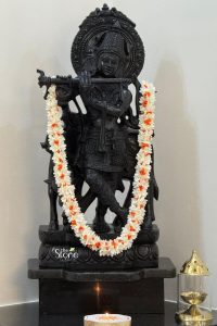 A detailed, hand-carved black stone Krishna statue stands as a serene centerpiece, reflecting the traditional artistry found in spiritual decor shops in Bangalore. This approximately 2-foot idol depicts Lord Krishna in the classic Tribhanga posture, gracefully playing his flute. Meticulously sculpted from dark, polished stone, the statue showcases intricate carvings on his ornate crown, jewelry, and robes, with a cow standing faithfully behind him—a symbol of his pastoral life. A long, festive garland made of white and orange flowers is draped around the deity's neck, providing a vibrant contrast against the dark stone. The statue is positioned on a sleek, matching black pedestal within a calm, domestic setting. In the foreground, a single lit diya casts a warm, flickering glow, while a small metallic decorative lamp sits to the side, enhancing the tranquil and meditative atmosphere. This arrangement illustrates how a beautifully crafted Krishna statue can serve as a focal point for home worship and aesthetic spiritual decor.