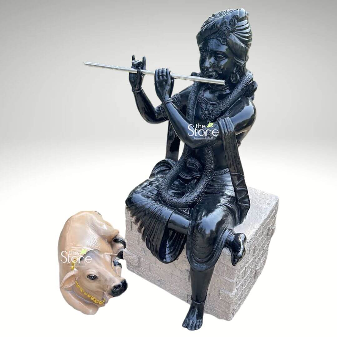 Lord Krishna with Cow Fibre Statue 4ft 8 An exquisitely crafted fibre statue of Lord Krishna Govinda, featuring a striking high-gloss black finish that radiates a divine and polished appearance. Shown from a three-quarter perspective, the sculpture depicts Lord Krishna in a relaxed and graceful seated pose atop a grey, stone-textured rectangular pedestal. He is captured in his iconic flute-playing gesture, holding a silver-toned flute with a serene and blissful facial expression. The deity is beautifully adorned with traditional Indian attire, including an intricately detailed turban, layered necklaces, and armlets. Resting faithfully at the base of the pedestal is a lifelike, light-brown sacred cow, adorned with a bright yellow beaded garland, symbolizing Krishna’s role as Govinda, the divine protector of animals. This durable and lightweight fibre sculpture is designed for both indoor and outdoor use, making it an excellent spiritual centerpiece for a home mandir, luxury foyer, or as outdoor garden decor. The contrast between the radiant black finish of the Krishna idol and the realistic tones of the cow creates a visually captivating focal point that invites an atmosphere of tranquility, devotion, and positive energy into any living space.