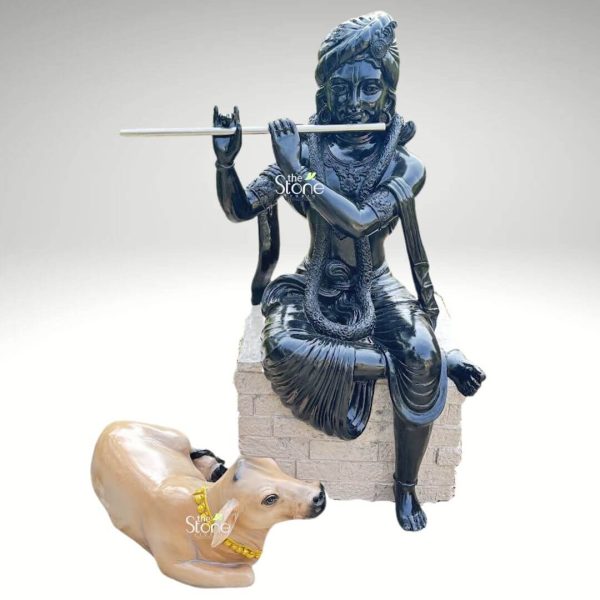 A serene and masterfully crafted fibre statue of Lord Krishna with a cow, featuring a striking high-gloss black finish that radiates a divine and polished appearance. The sculpture depicts Lord Krishna in a relaxed and graceful seated pose atop a grey, stone-textured rectangular pedestal. His hands are positioned delicately as he plays a long, silver-toned flute, his facial expression reflecting peace and spiritual bliss. The deity is beautifully adorned with traditional Indian attire, including an intricately detailed turban, layered necklaces, armlets, and a dhoti with realistic fabric folds. Resting faithfully at the base of the pedestal is a lifelike, light-brown cow with a bright yellow beaded garland around its neck, symbolizing Krishna’s role as the divine protector of animals. This durable and lightweight fibre sculpture is designed for both indoor and outdoor use, making it an excellent spiritual centerpiece for a home temple, commercial foyer, or as outdoor garden decor. The combination of the deep black Krishna idol and the naturally colored cow creates a visually captivating focal point that invites an atmosphere of tranquility, devotion, and positive energy.