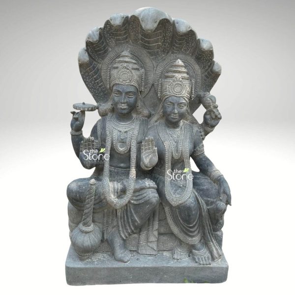 An exquisitely hand-carved Lord Narayan Lakshmi black granite statue, capturing the divine couple in a serene seated posture. This blackstone statue depicts Lord Vishnu (Narayan) and Goddess Lakshmi seated together, framed by the majestic multi-headed hoods of the serpent Sheshanaga, which forms a protective and ornate canopy over them. Lord Narayan is shown with four arms, holding the sacred Sudarshana Chakra (discus) and the Panchajanya Shankha (conch), while his lower right hand is raised in the Abhaya Mudra, a gesture of fearlessness and blessing. Resting beside him is his powerful mace, the Gada. Goddess Lakshmi, the deity of wealth and prosperity, sits gracefully by his side, also offering a blessing. Both figures are adorned with towering, tiered crowns (mukuts), intricate necklaces, and traditional garments featuring finely rendered fabric folds. The sculpture is masterfully crafted from a single block of natural black granite, showcasing a refined matte finish that highlights the artisan's attention to detail. This durable and weather-resistant Narayan Lakshmi murti serves as a profound spiritual centerpiece for a home mandir, meditation room, or an elegant garden sanctuary, symbolizing divine harmony, protection, and abundance.