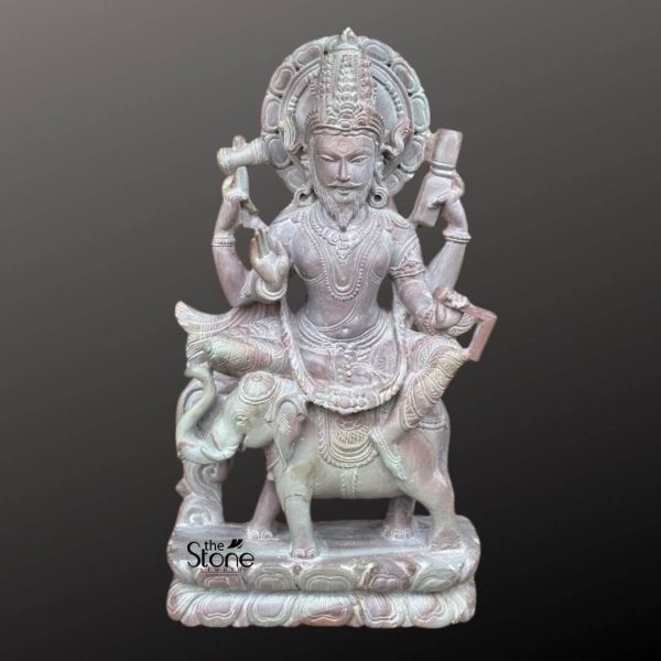 An intricately hand-carved stone statue of Lord Vishwakarma, the divine architect of the universe, is depicted in a majestic seated posture atop his vahana, a finely detailed elephant. The idol is crafted from high-quality natural stone with an earthy pinkish and grey matte finish, showcasing masterful craftsmanship throughout. The four-armed deity holds traditional tools of creation and spiritual wisdom, including a measuring scale, a water pot, and sacred texts, while one hand is raised in the Abhaya Mudra, a sacred gesture of protection and blessing. Lord Vishwakarma is adorned with an elaborate multi-tiered crown, layered ornamental necklaces, and a decorative circular halo that frames his serene, bearded face. The entire sculpture rests on a solid, integrated rectangular base with subtle carvings. This traditional stone idol serves as a powerful symbol of creativity and engineering, making it an ideal spiritual centerpiece for architectural studios, workshops, or home temples.