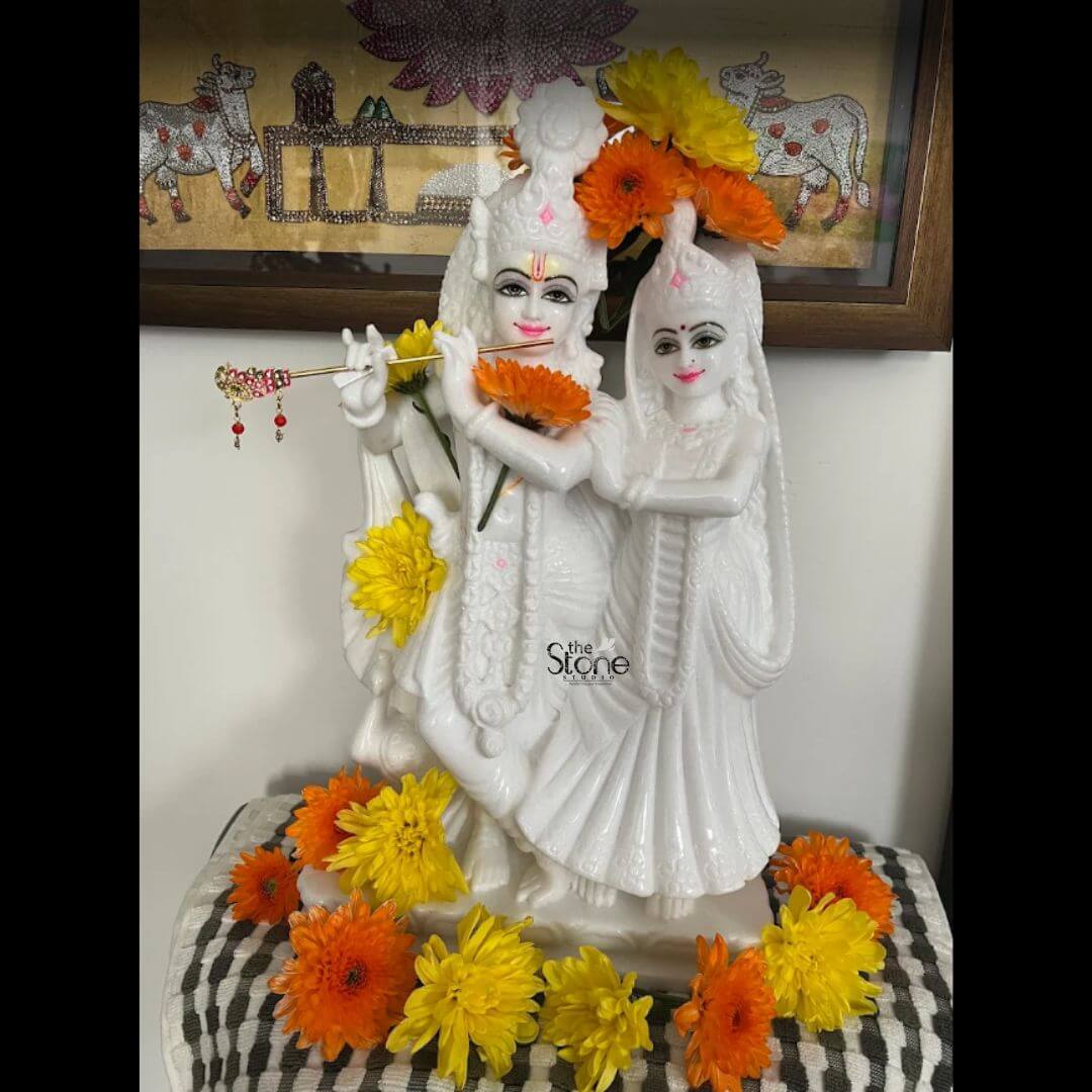 18 inch Marble Radha Krishna Statue 6 A radiant marble Radha Krishna statue adorned with fresh orange and yellow flowers is presented in a close-up front-facing view captivatingly displayed in an indoor setting. The statue beautifully depicts Lord Krishna playing his flute while Goddess Radha stands lovingly by his side their forms sculpted from lustrous white marble that gleams softly. This exquisite marble Radha Krishna statue is a vibrant and spiritual centerpiece perfect for a home temple or decor in Melbourne symbolizing divine love and harmony. Both Radha and Krishna are intricately detailed with delicate facial features painted to enhance their expressions: Krishna has rosy lips, a bindi and gentle eyes while Radha shares similar delicate features both wearing serene smiles. Krishna is adorned with a small crown and holds a beautifully embellished flute to his lips while Radha is depicted in flowing drapery with her hands gracefully positioned. Fresh orange and yellow marigold-like flowers are reverently placed around their necks at their feet and tucked into their crowns adding bursts of color and traditional vibrancy to the pristine white marble. In the background a framed artwork featuring traditional Indian art with cows further enhances the cultural context. This marble Radha Krishna statue is a profound representation of divine love and spiritual devotion making it a cherished addition to any home in Melbourne. The pure white marble evokes purity and serenity while the vibrant floral offerings signify reverence and celebration. The detailed craftsmanship and the inherent beauty of the marble combine to create a deeply meaningful and aesthetically pleasing artifact fostering an atmosphere of peace love and spiritual connection within any living space.