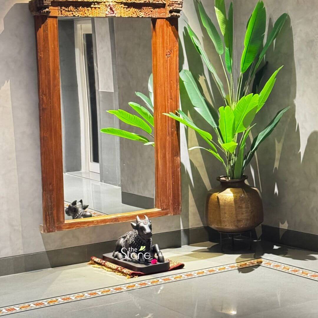 A serene 2-foot tall Lord Nandi statue, hand-carved from natural black stone, serves as a spiritual focal point in a modern foyer in Vijayawada. The blackstone Nandi idol is depicted in a traditional recumbent pose, resting on a small red-and-gold patterned rug. The sculpture is artfully placed in a minimalist corner featuring a large, full-length mirror with a rustic dark-wood antique frame. Flanking the setup is a lush green tropical plant in a massive, aged brass pot, set against contemporary grey concrete-textured walls. Polished grey marble floors with an intricate tile border reflect the warm ambient lighting which casts a soft shadow of the Nandi statue and its reflection in the mirror. This sophisticated interior design choice highlights the blend of ancient Indian heritage and modern home decor creating a peaceful and auspicious atmosphere in a Vijayawada residence.