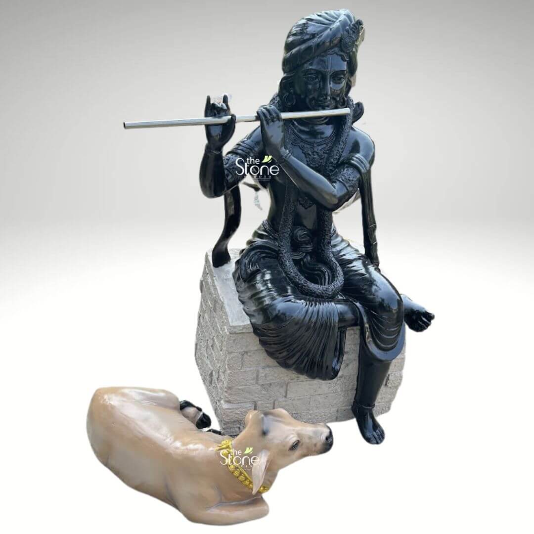 Lord Krishna with Cow Fibre Statue 4ft 4 An exquisitely crafted premium black fibre Lord Krishna with cow statue, showcasing a striking high-gloss finish that radiates a divine and polished appearance. The fibre statue depicts Lord Krishna in a relaxed and graceful seated pose atop a light grey, stone-textured rectangular pedestal. He is captured in his iconic musical gesture, holding a sleek silver-toned flute to his lips with a serene and blissful facial expression. The deity is beautifully adorned with intricate traditional Indian attire, including a detailed turban, layered necklaces, armlets, and a dhoti with realistic fabric folds. Resting faithfully at the base of the pedestal is a lifelike, light-brown sacred cow, adorned with a bright yellow beaded garland, symbolizing Krishna’s role as the divine protector of animals. This durable and lightweight fibre sculpture is designed for both interior and exterior use, making it an excellent spiritual centerpiece for a home mandir, a luxury foyer, or as sophisticated outdoor garden decor. The sharp contrast between the radiant black finish of the Krishna idol and the realistic tones of the cow creates a visually captivating focal point that invites an atmosphere of tranquility, devotion, and positive energy into any living space.