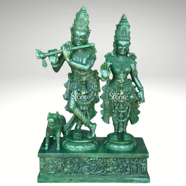 An exquisite 3-foot tall Radha Krishna green stone statue, masterfully hand-carved to capture the divine couple in a serene and graceful standing pose. Lord Krishna is depicted in his iconic cross-legged posture, delicately playing the flute, while Radha stands beside him with her right hand raised in the Abhaya Mudra of blessing. Both figures are adorned with towering, intricately detailed crowns, layered necklaces, and traditional ornaments that showcase fine craftsmanship. A small calf, representing innocence and devotion, stands faithfully near Krishna's feet. The entire sculpture is made of polished green stone with a rich emerald luster. They stand on individual lotus bases atop a large rectangular pedestal featuring high-relief carvings of smaller figures in various devotional poses. This 3ft green stone Radha Krishna idol is an ideal spiritual centerpiece for a home mandir, garden, or grand living space, radiating peace and divine love.